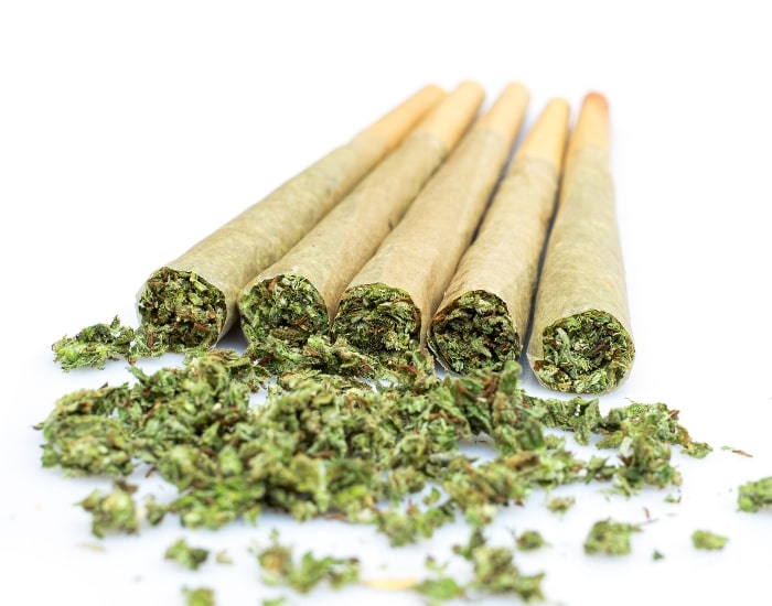 Five rolled cannabis joints with loose marijuana on white background for smoking use.