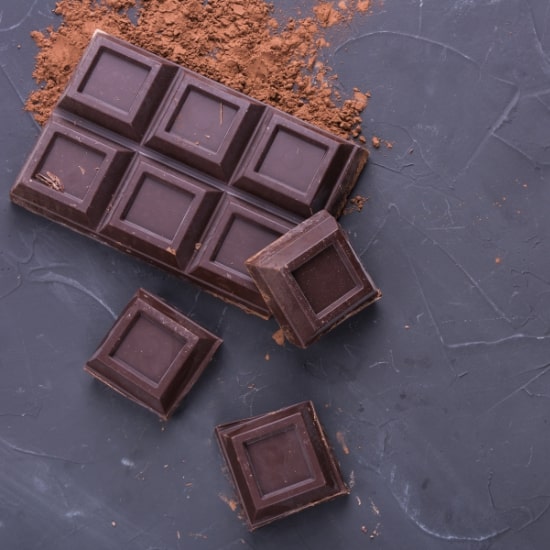 Dark chocolate bar with broken pieces and cocoa powder on slate background.