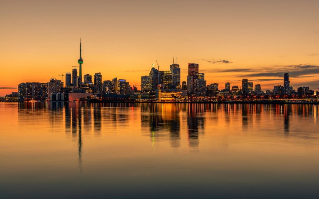 Toronto skyline at sunset with CN Tower reflected on calm waterfront.