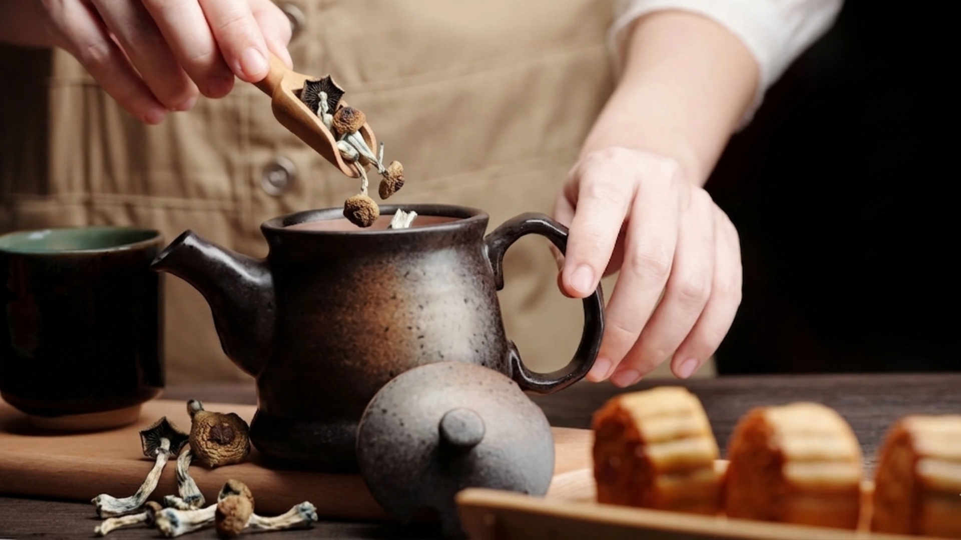 A person using a wooden scoop to add dried psilocybin mushrooms into a dark ceramic teapot.