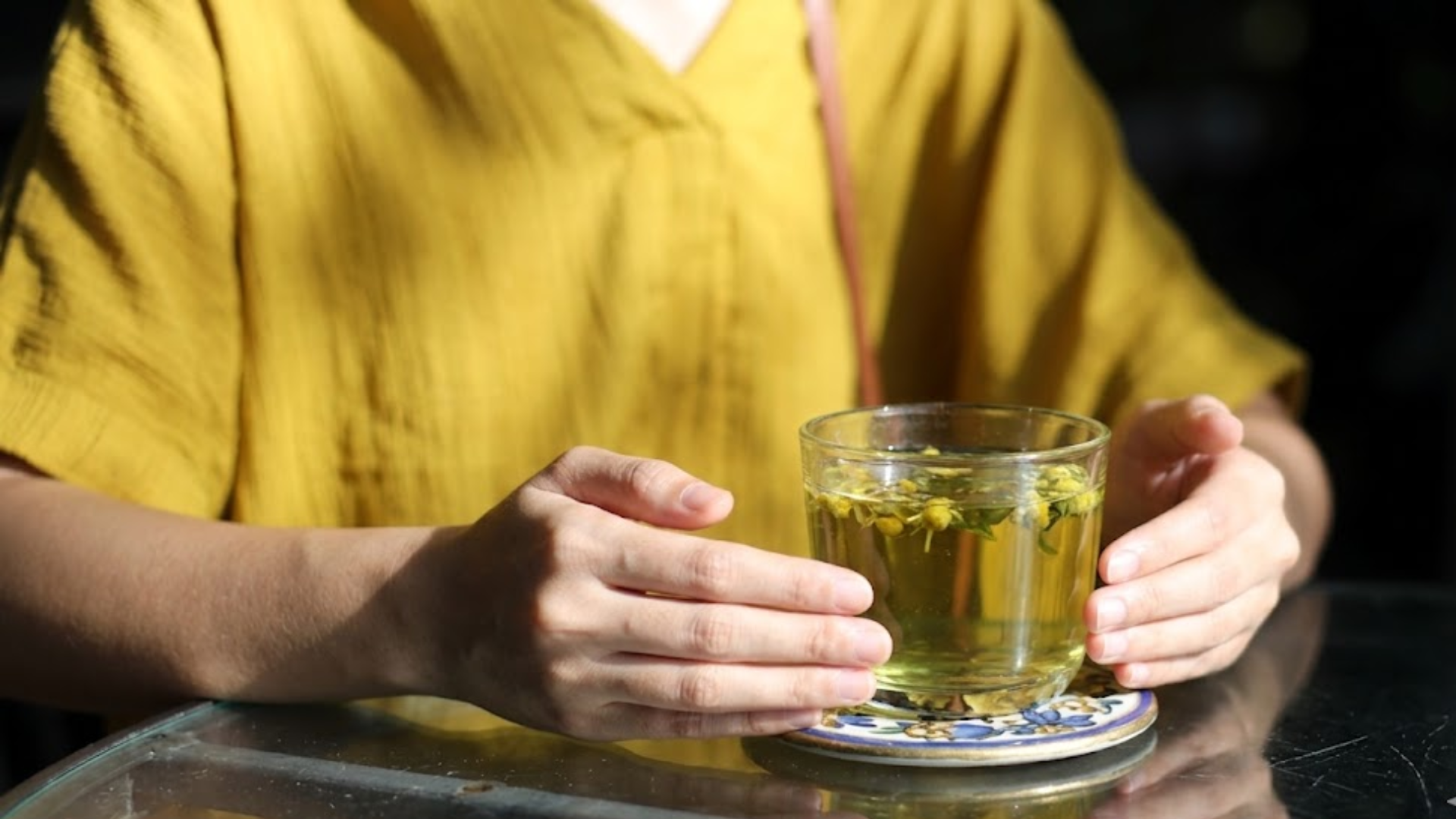 A person in a yellow shirt sits at a table with a glass of green tea in a sunlit setting.